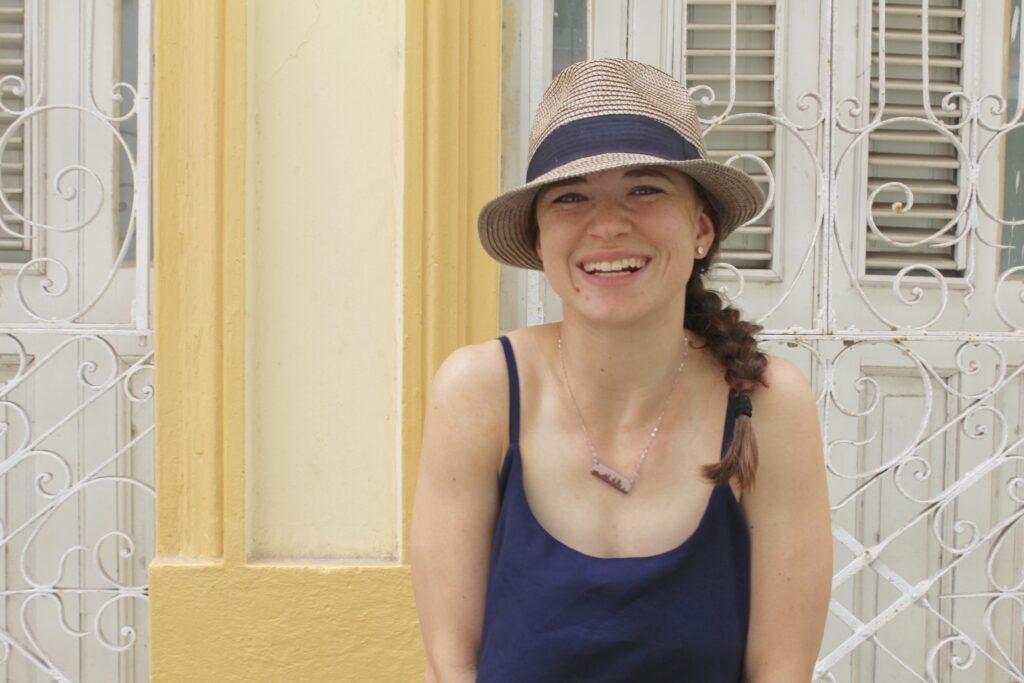Girl with fedora and braid smiling in front of a yellow, colonial style building