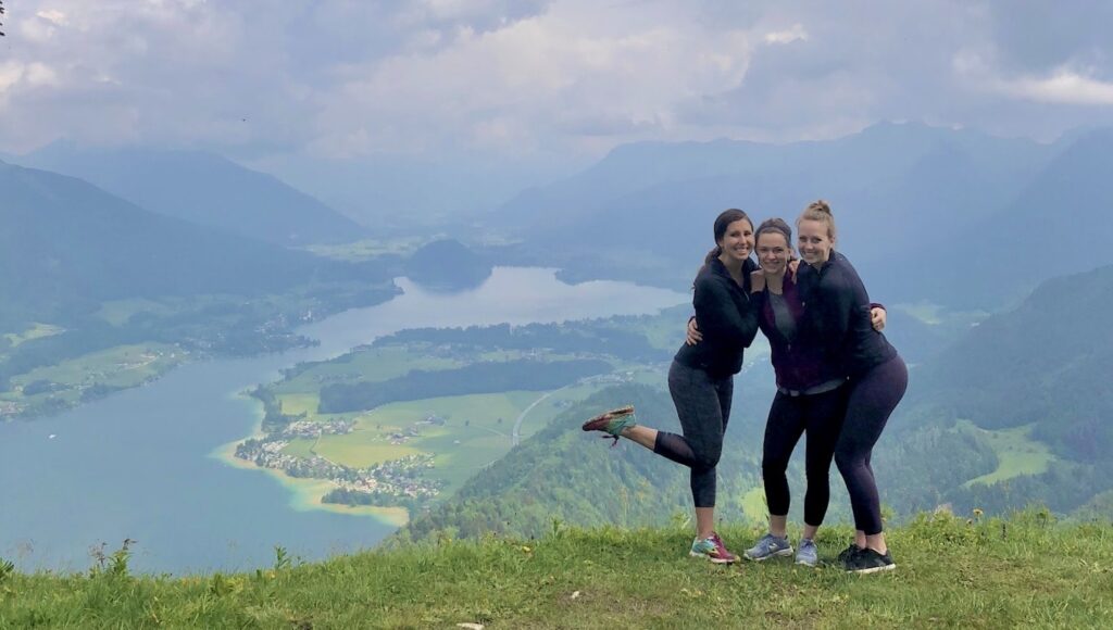 Three women smiling and embracing each other while standing on a hilltop with a panoramic view of a valley, including a winding river and distant mountains under a cloudy sky.