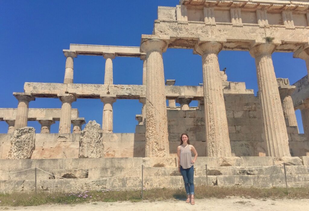 A woman standing in front of the ruins of an ancient Greek temple with large columns and a bright blue sky in the background.