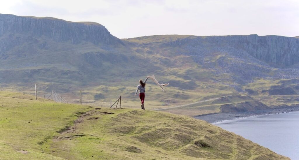 A woman running on a grassy hill with a scarf trailing behind her. The background features a rugged, mountainous landscape near the coast.