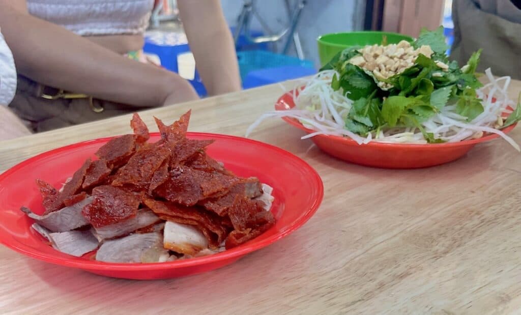 Two red bowls on a table. In one bowl is stacked dried meat and in the other is shreds of light green papaya topped with greens and roasted peanuts.