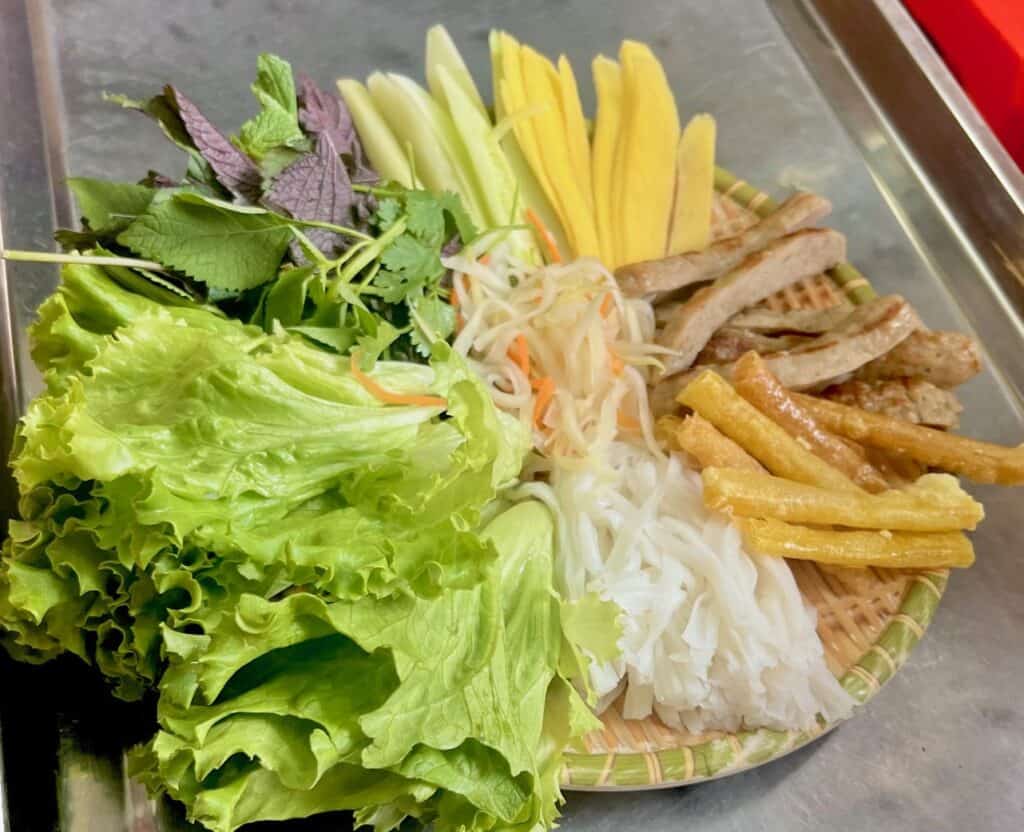 A plate containing lettuce, noodles, sausages, mangoes, mint, cilantro, and sliced cucumber.