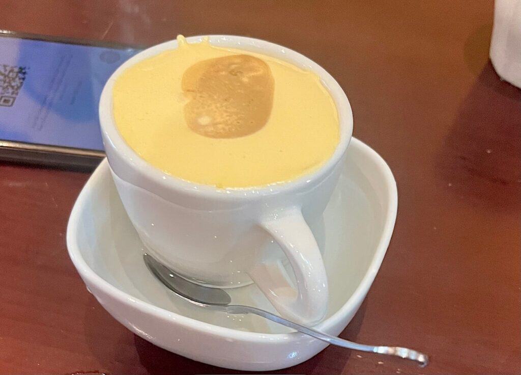 A small coffee cup on a saucer with a spoon with a thick creamy custard at the top of the cup.