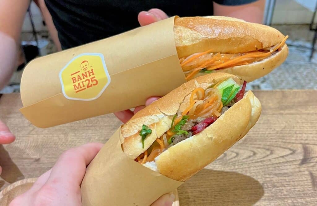 Two hands holding matching sandwiches. The sandwiches are made of baguettes with carrots, cilantro, and meat. The wrapper says Bahn Mi 25 on the sandwich.