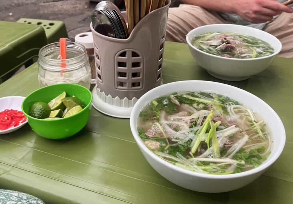 Two bowls of pho on a green plastic table. On the table is also limes, chiles, and chopsticks.