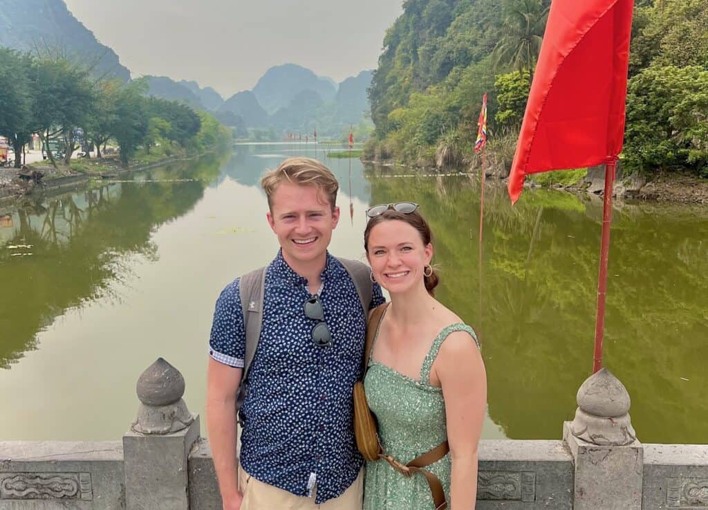 A smiling couple standing on a bridge overlooking a serene river  in Ninh Binh with lush green trees and mountains in the background, adorned with red flags on the sides.