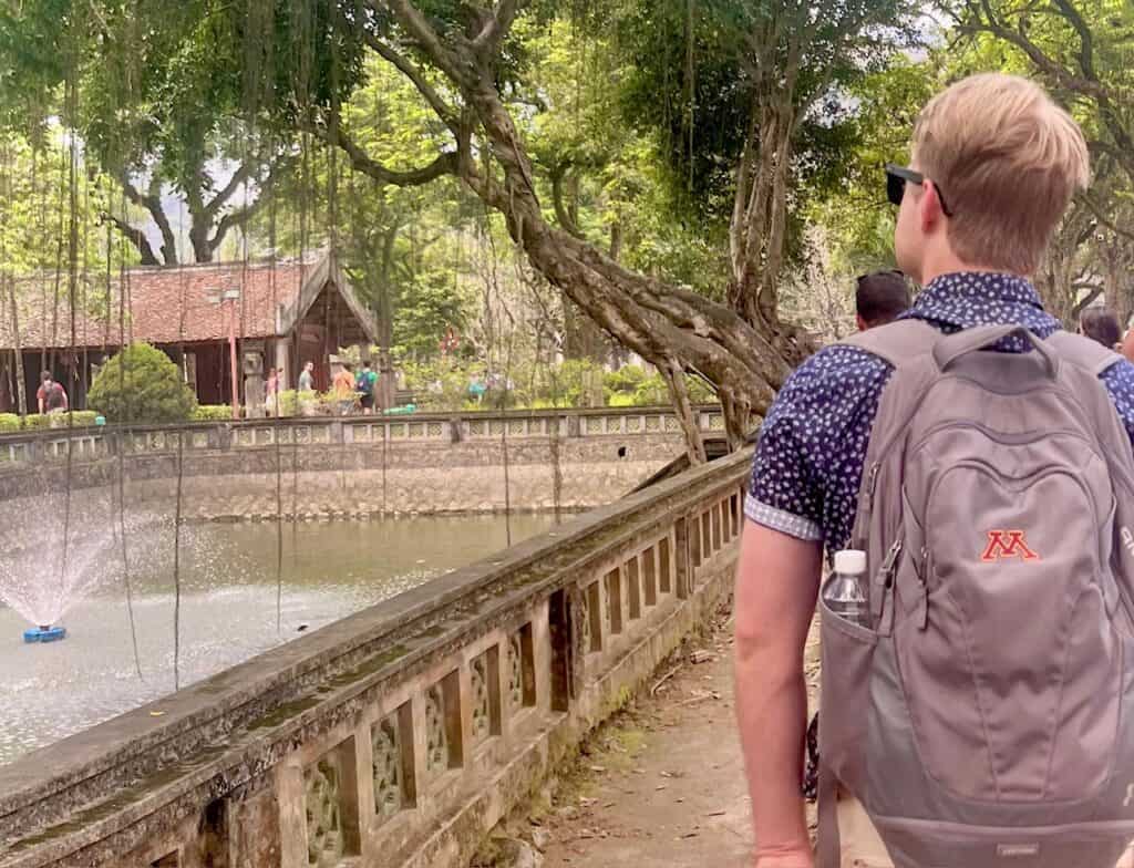 A person with a backpack walking along a path next to a stone railing, with a fountain spraying water in a pond and a traditional wooden building visible in the background.