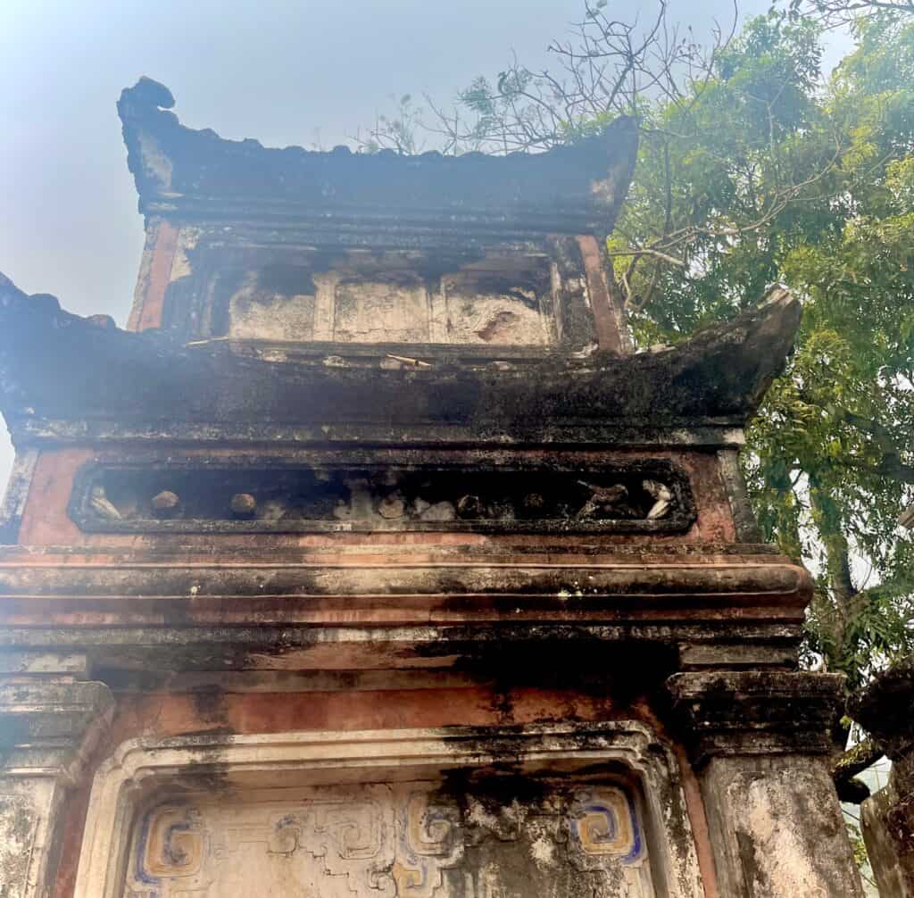 A close-up view of an old, weathered stone structure with ornate carvings, partially covered by tree branches and foliage on a day trip from Hanoi to Ninh Binh.