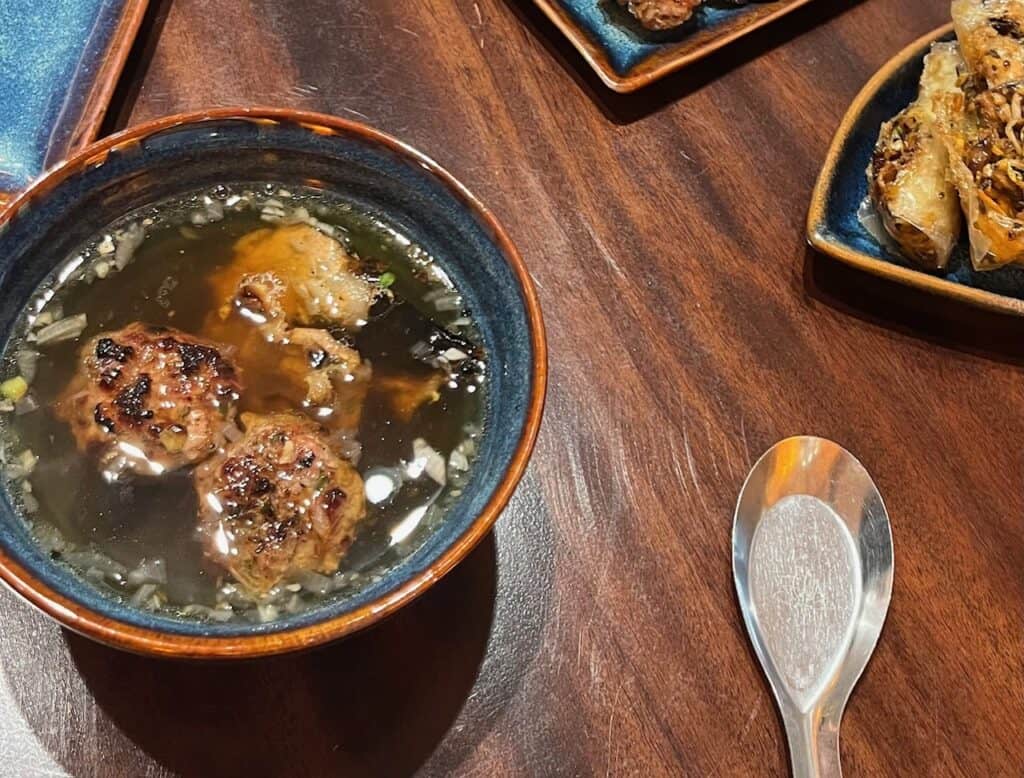 A bowl on a table with a large metal spoon next to it. The contents of the bowl are broth and meatballs.