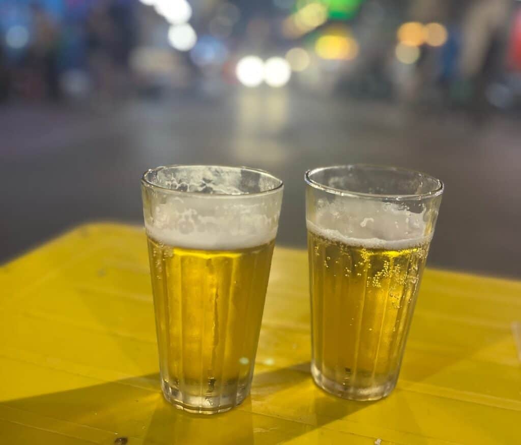 Two glasses of light beer with foam on the top. The glasses are sitting on a plastic yellow table with a busy street in the distance.