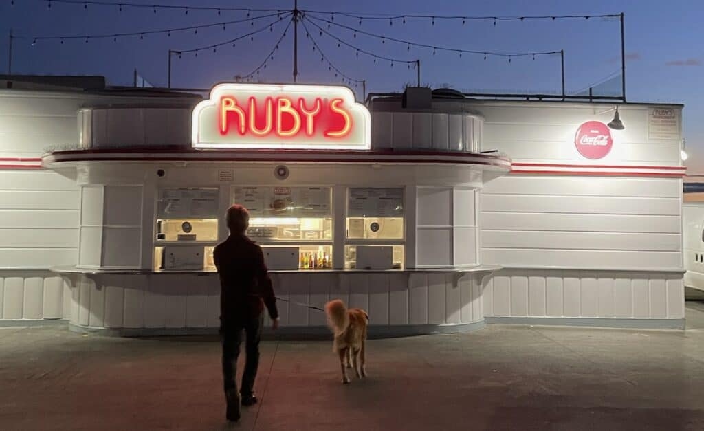 Night scene of a person and a dog standing in front of a retro-style diner called Ruby's, illuminated by a bright neon sign. The background includes string lights and a Coca-Cola sign.