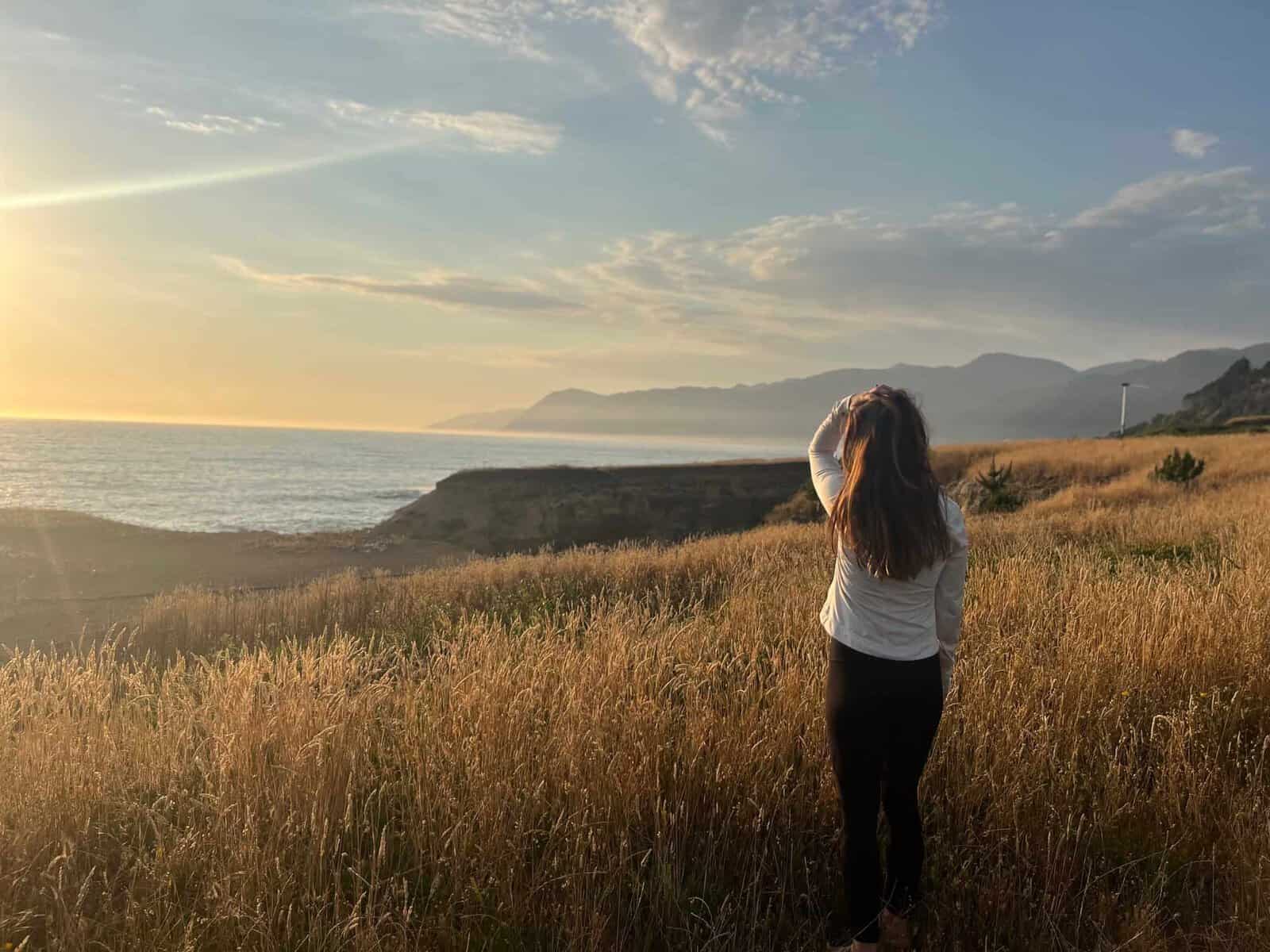 Woman in white long sleeve with hand in her hair overlooks the golden wheat fields with the ocean and mountains of Shelter Cove in the distance.