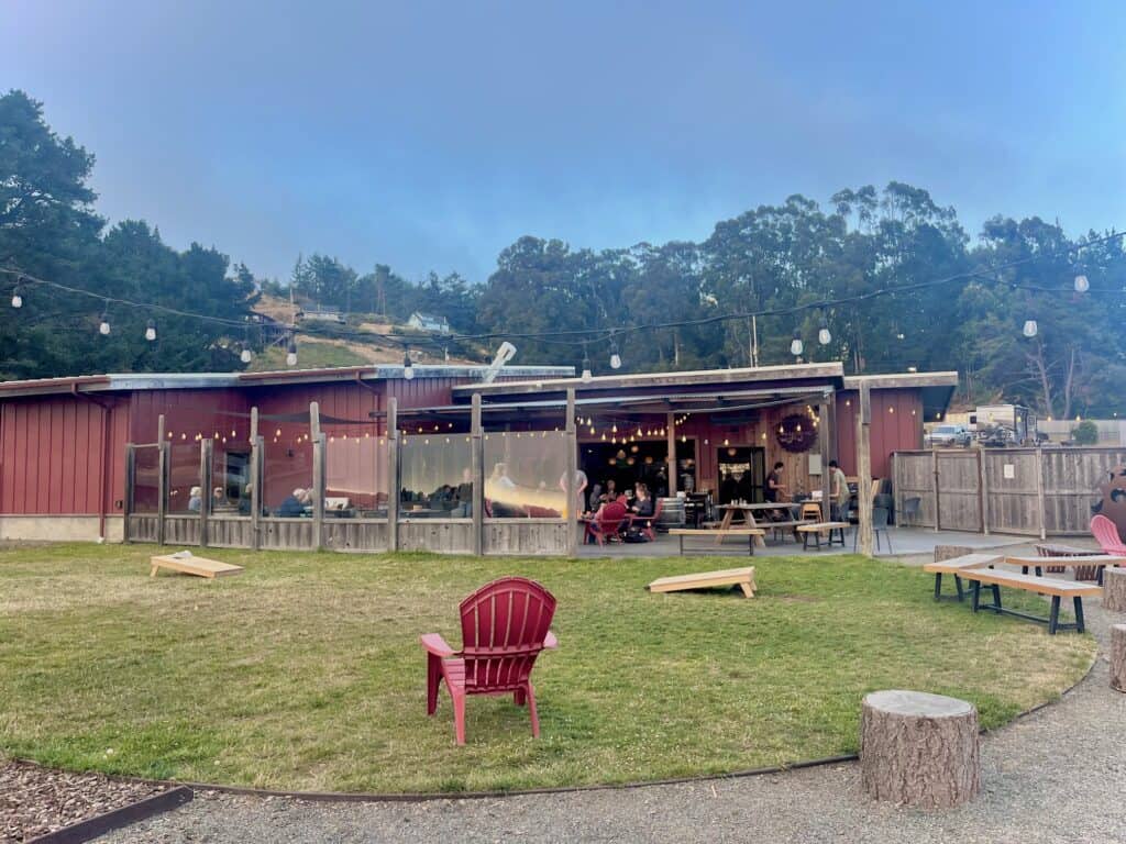 A red brewery with string lights surrounding the  outdoor patio. Corn hole boards are set up in the grassy area.