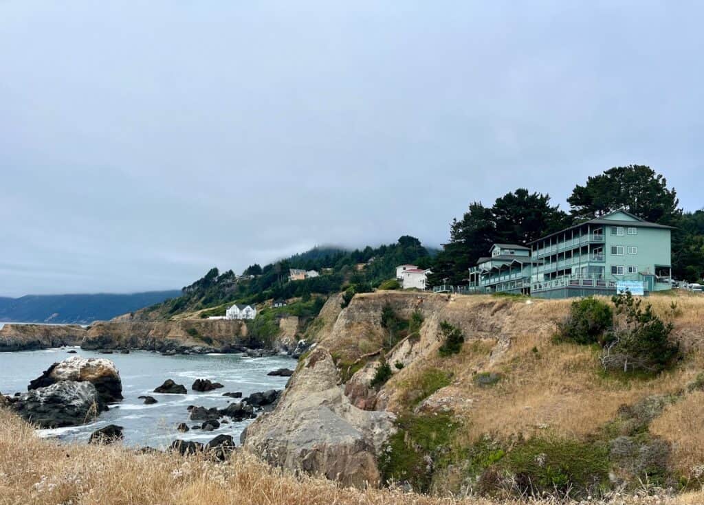 Cliffside view with houses and the Inn of the Lost Coast overlooking a rocky beach. The sky is overcast, and the area is surrounded by green vegetation and dry grass.