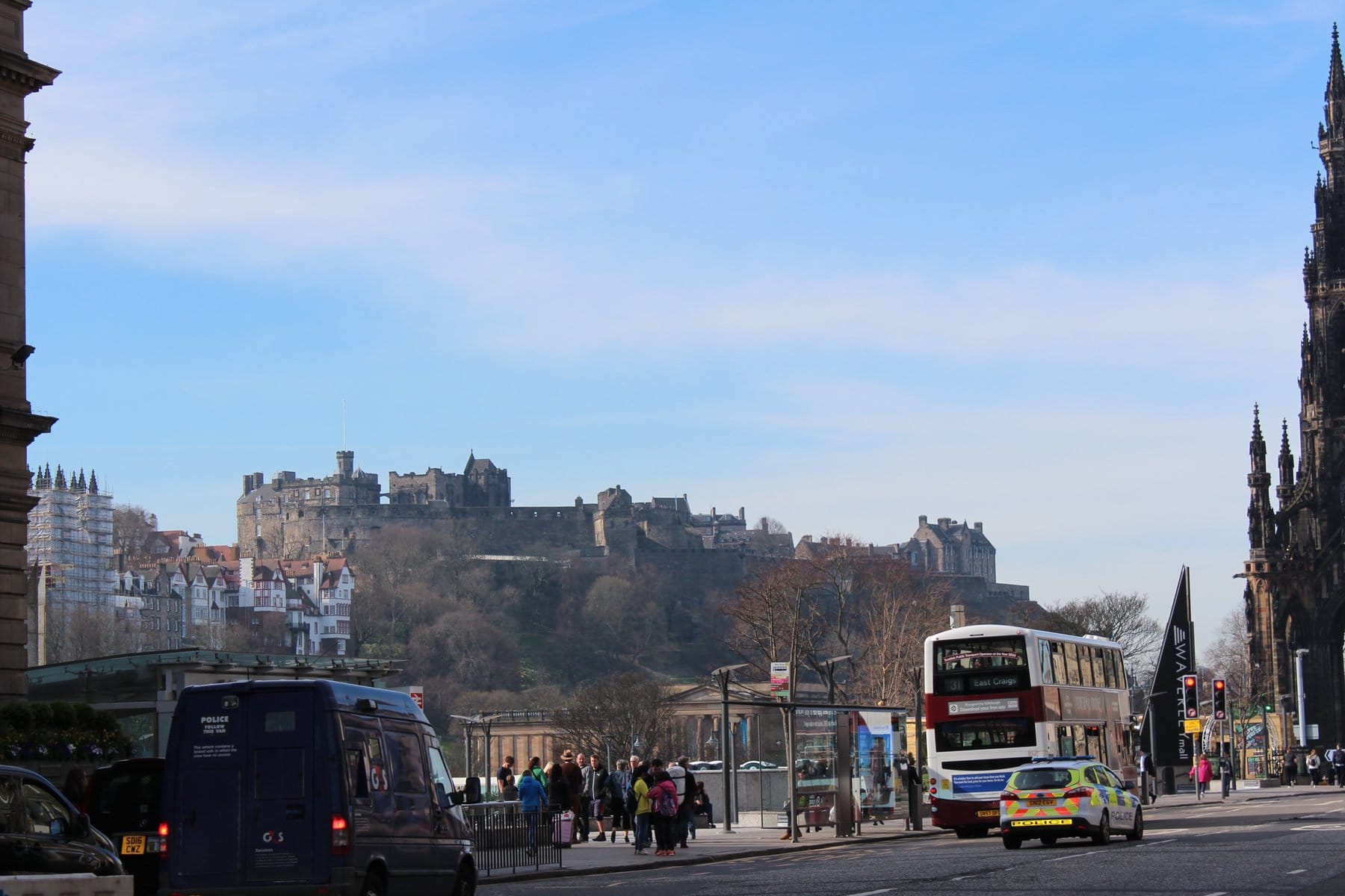 A street view featuring a police van, a double-decker bus, and pedestrians, with the historic Edinburgh Castle in the background on a sunny day. A great spot to visit with 1 day in Edinburgh.