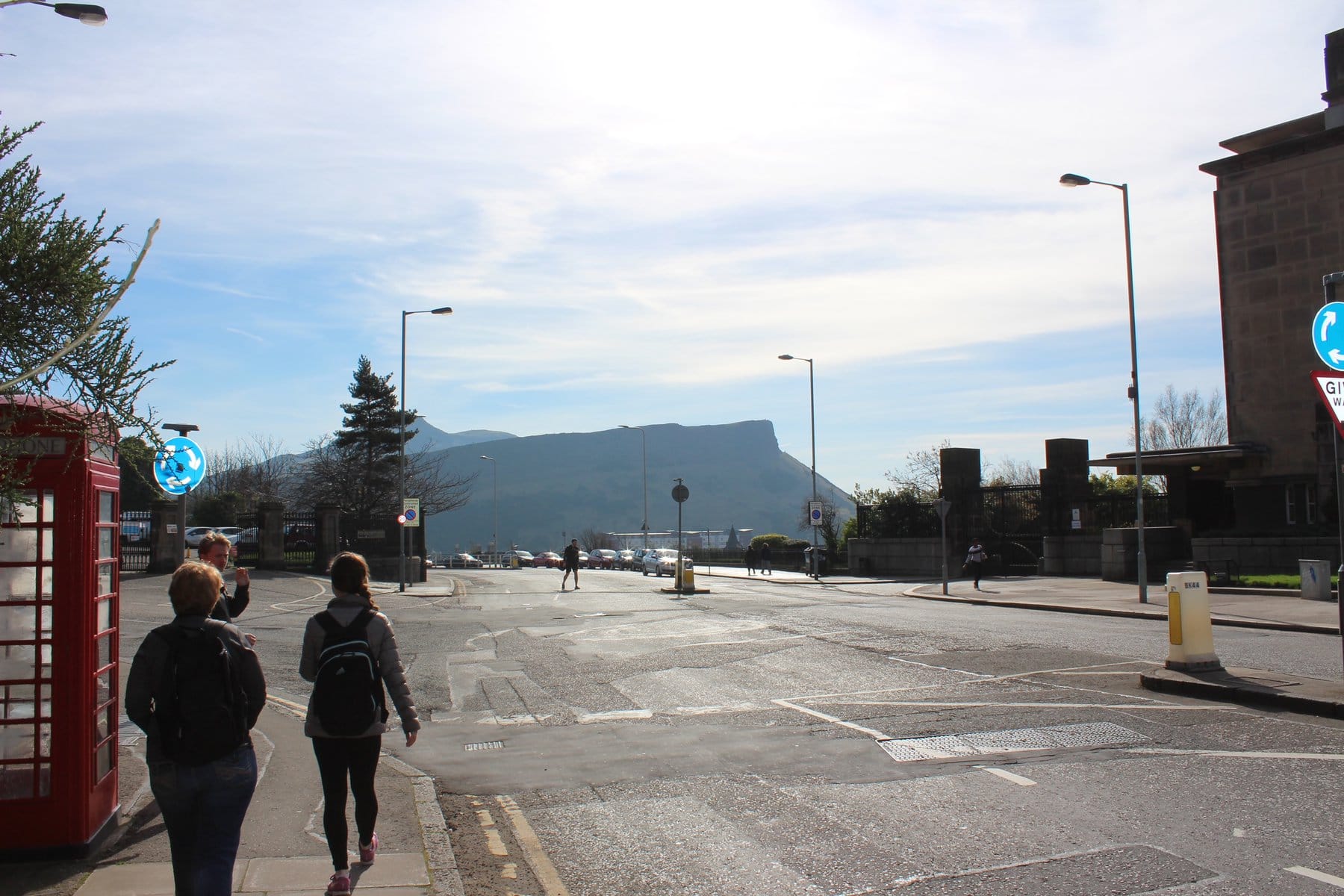 Street scene in Edinburgh with people walking on the sidewalk, a red phone booth on the left, and the silhouette of Arthur's Seat in the background under a bright sky.