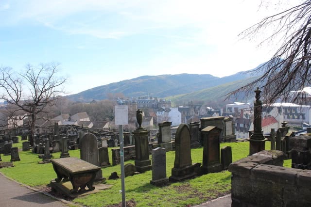 A scenic view of an old cemetery with various tombstones and monuments, set against a backdrop of rolling hills and a small town. The clear sky and the distant mountains add to the tranquil atmosphere of the scene.