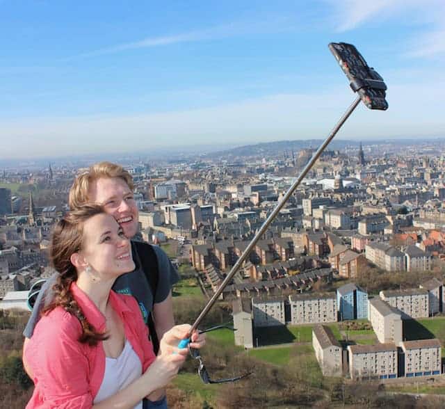  A young couple taking a selfie with a selfie stick, overlooking a sprawling cityscape on a clear day in Edinburgh. The city below features a mix of residential and commercial buildings.