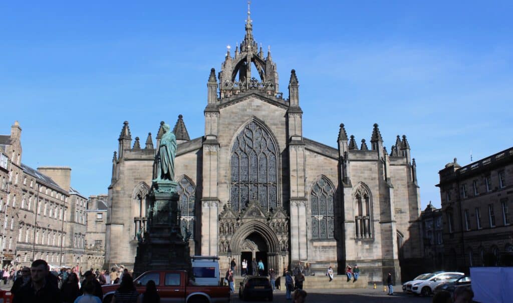 A bustling square featuring a large Gothic-style St Giles Cathedral with intricate stained glass windows and spires. A statue stands prominently in front, with people walking and cars parked nearby under a bright blue sky.