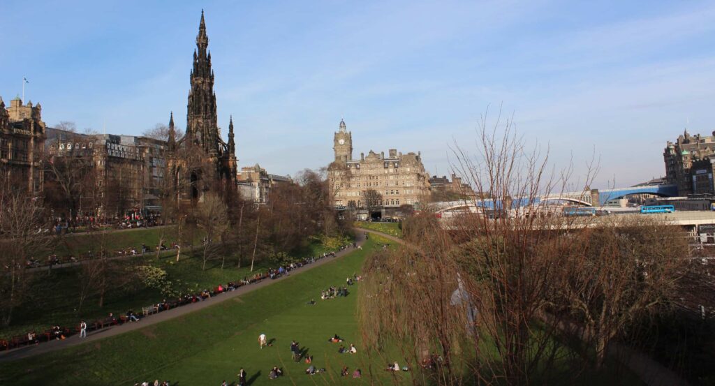 A wide view of a lush green park filled with people relaxing and walking, surrounded by historical buildings including a tall gothic spire and an elegant clock tower. 