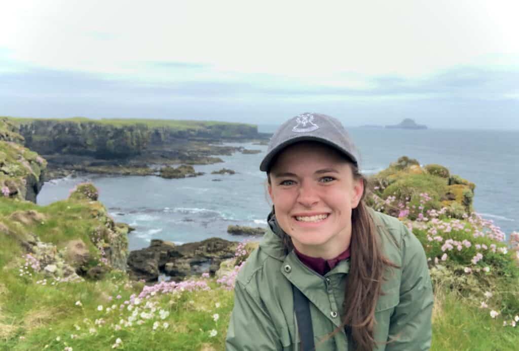 A woman smiling in a green jacket and baseball cap, standing on a cliff with the ocean and rocky coastline in the background, surrounded by pink flowers.