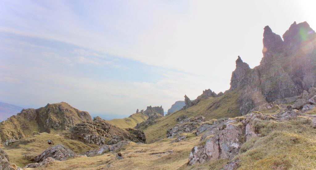 A dramatic mountain landscape at Isle of Skye in 1 day with rugged peaks and rocky terrain under a partly cloudy sky. The scene shows steep, jagged cliffs and grassy areas between the rocks.