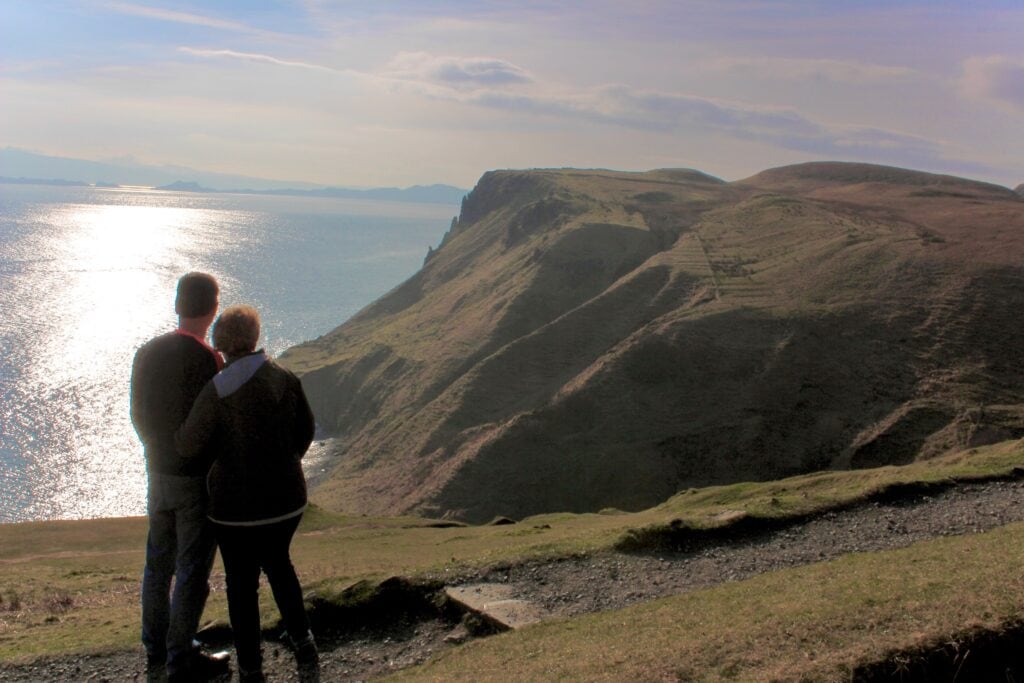 Two people stand close together, gazing out over a cliffside view of the Isle of Skye in 1 day with the sun reflecting off the ocean. The rolling hills and distant coastline add depth to the scenic landscape.