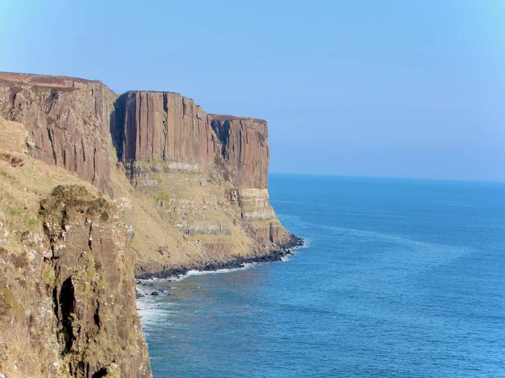 Rocky cliffs rise steeply from the sea, with layered rock formations visible. The blue ocean stretches into the horizon under a clear sky in 1 day in Isle of Skye.