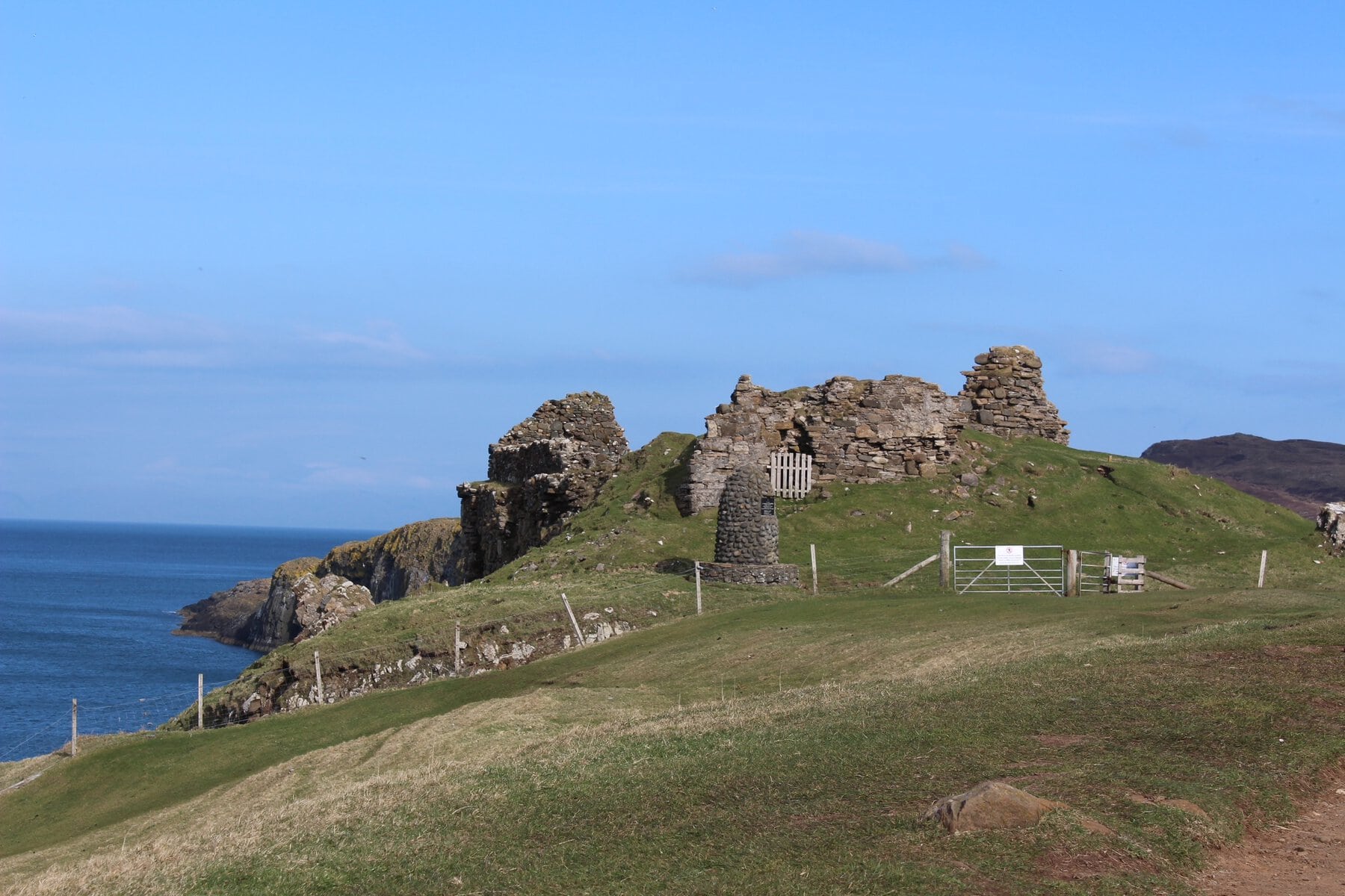 Ruins of a stone structure stand on a grassy hill overlooking the ocean. The blue sky and sea provide a serene backdrop to the ancient remnants.