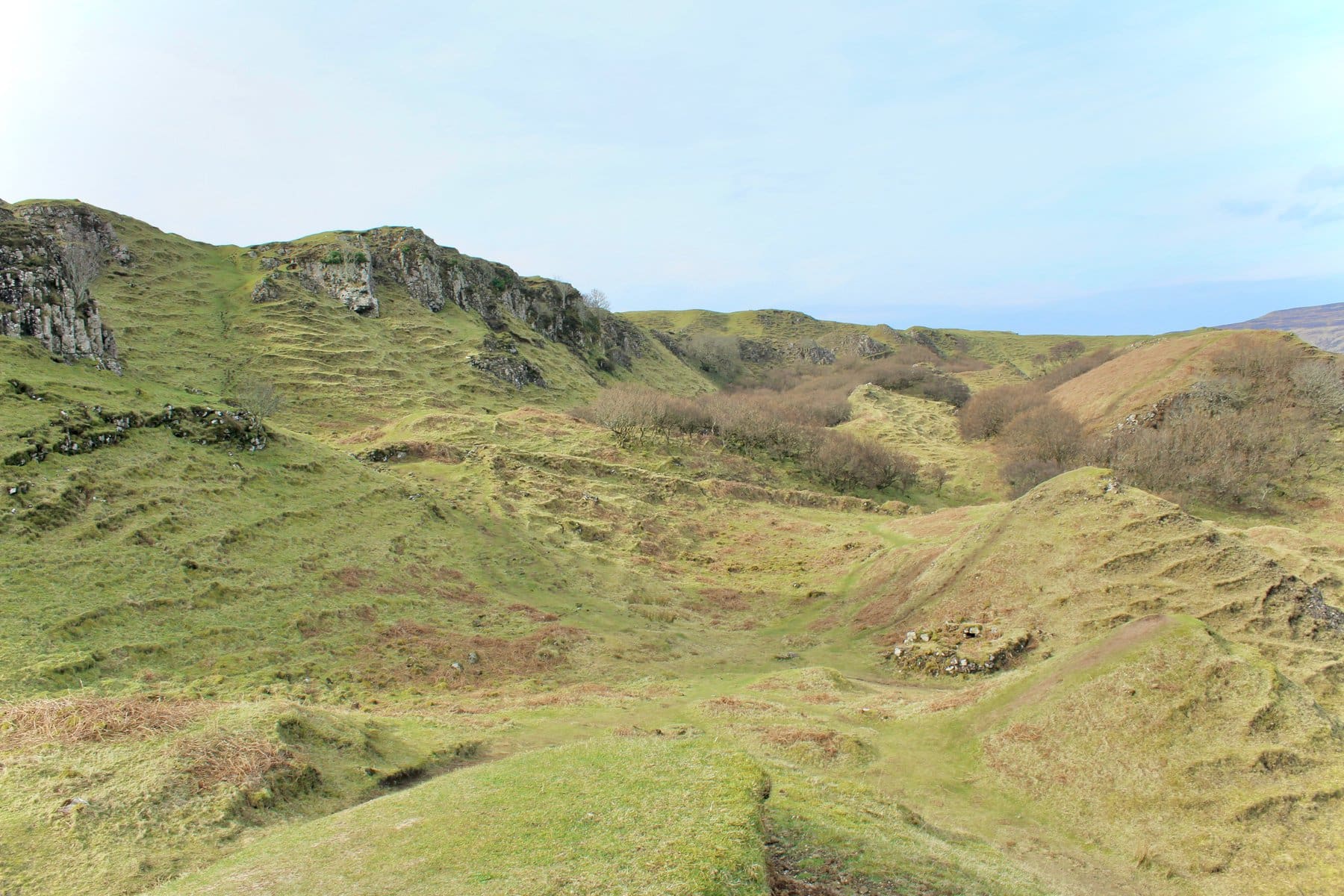  A wide shot of a grassy, hilly landscape with rocky outcrops and patches of shrubs under a light blue sky.