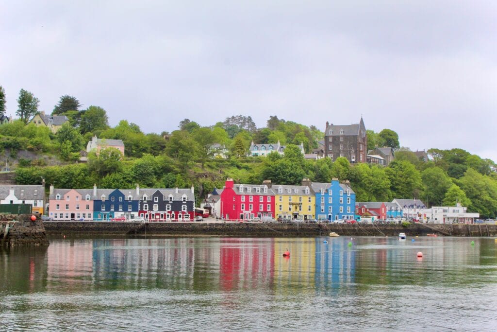 A picturesque row of colorful houses along the waterfront in Tobermory, Isle of Mull, Scotland, with lush greenery and a church in the background.