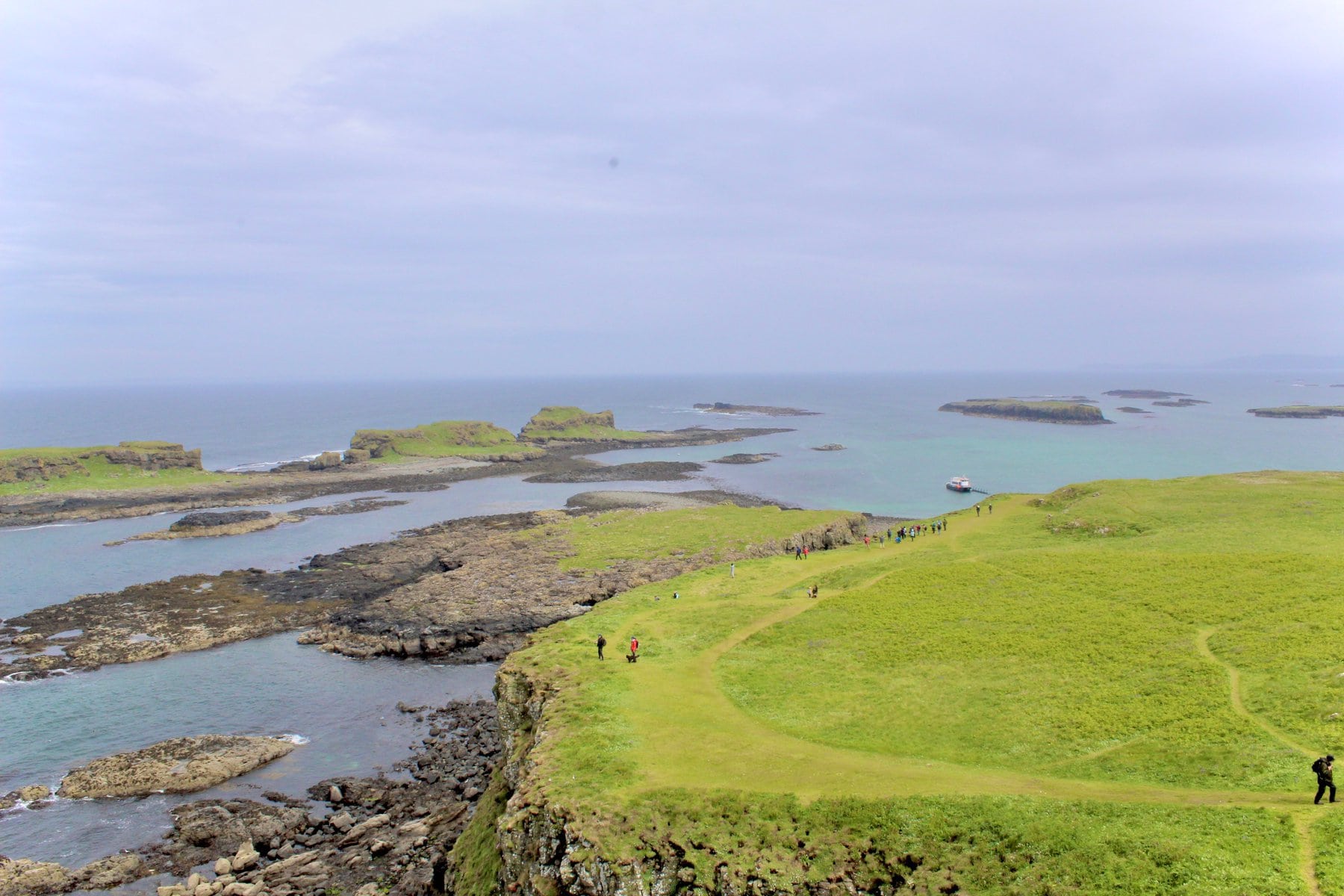 An expansive view of a rocky coastline with small islands in the distance, green grassy paths, and several people walking near the edge of the cliffs. A boat is docked at a distant shore.