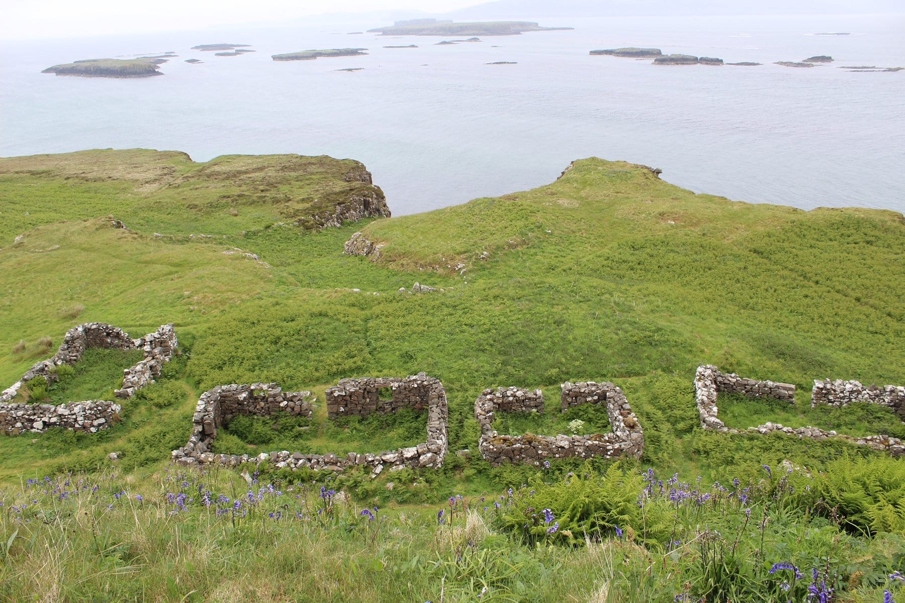 Stone ruins on a grassy hill overlooking the ocean, with scattered small islands in the distance and purple wildflowers in the foreground.