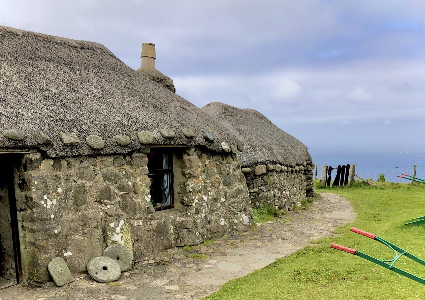 A traditional stone cottage at the Skye Museum of Island Life with a thatched roof stands in a grassy yard with a stone path leading towards the ocean. To the right, a green and red vintage cart is placed on the lawn under a cloudy sky.