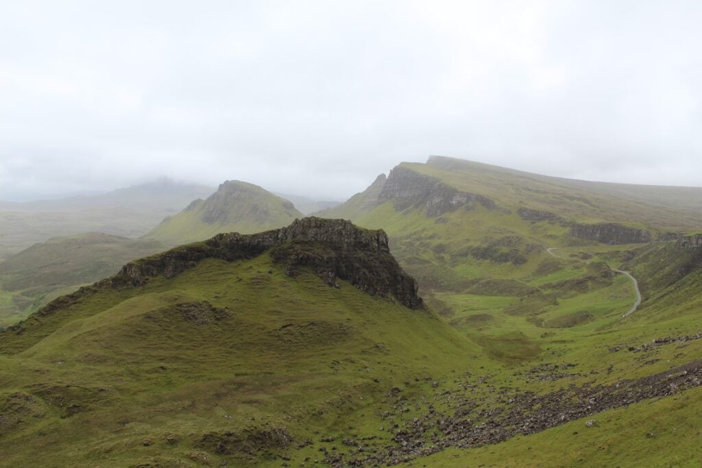 A panoramic view of lush green hills and mountains under a cloudy sky in the Isle of Skye. The landscape features rolling hills, rocky outcrops, and a winding path leading through the valley.