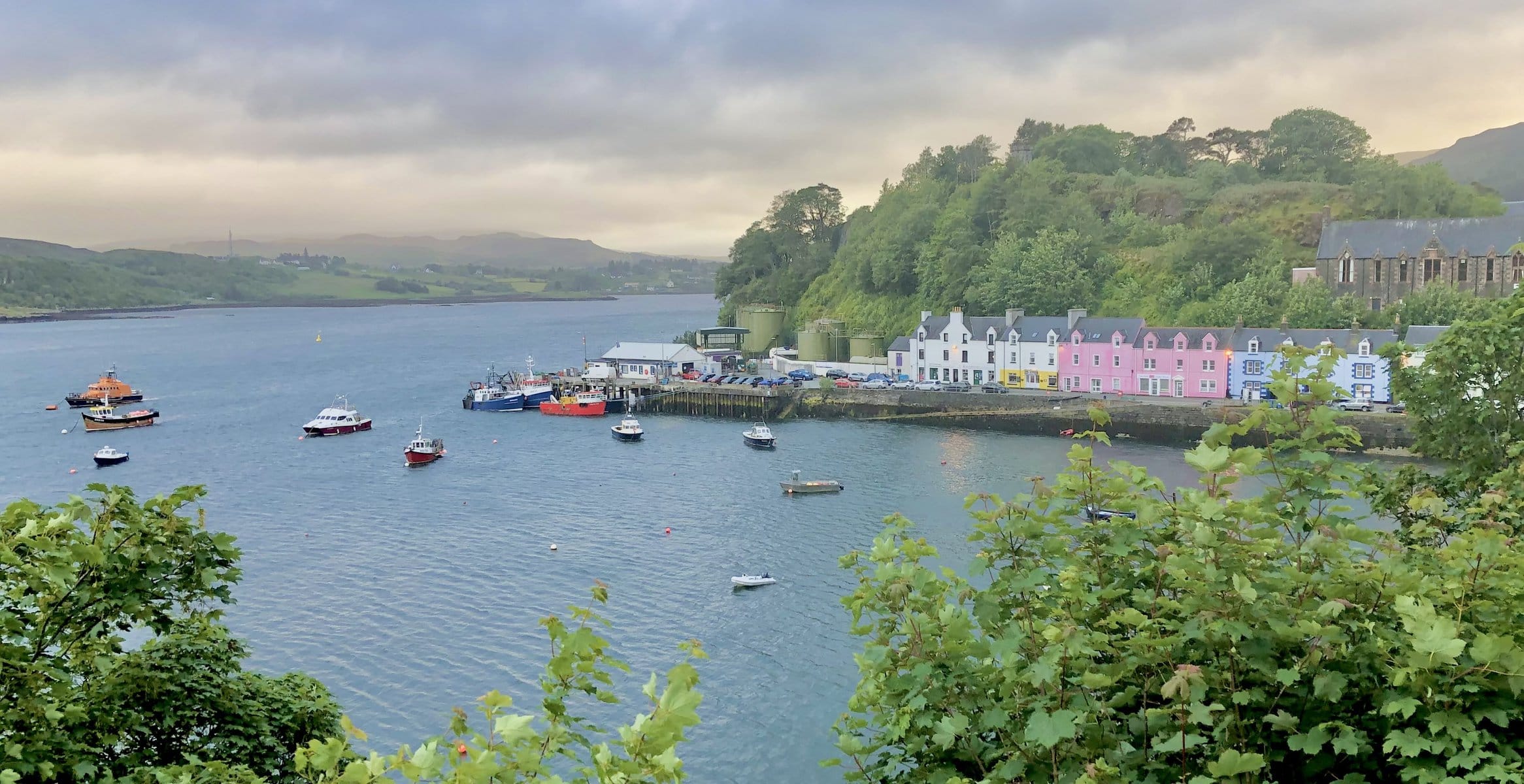 Pastel colored buildings lining Portree Harbor in Isle of Skye with multiple boats floating as the sun sets after the rain