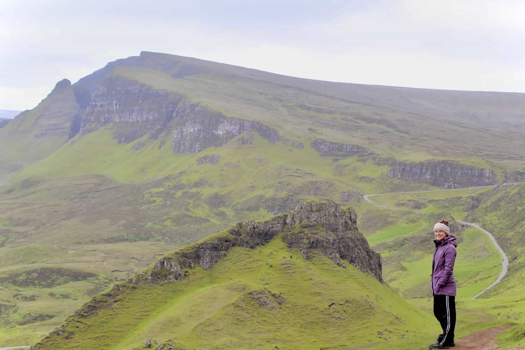 A woman in a purple jacket and a beige headband stands on a green hillside with a scenic view of rolling hills, rocky cliffs, and a winding road in the background.