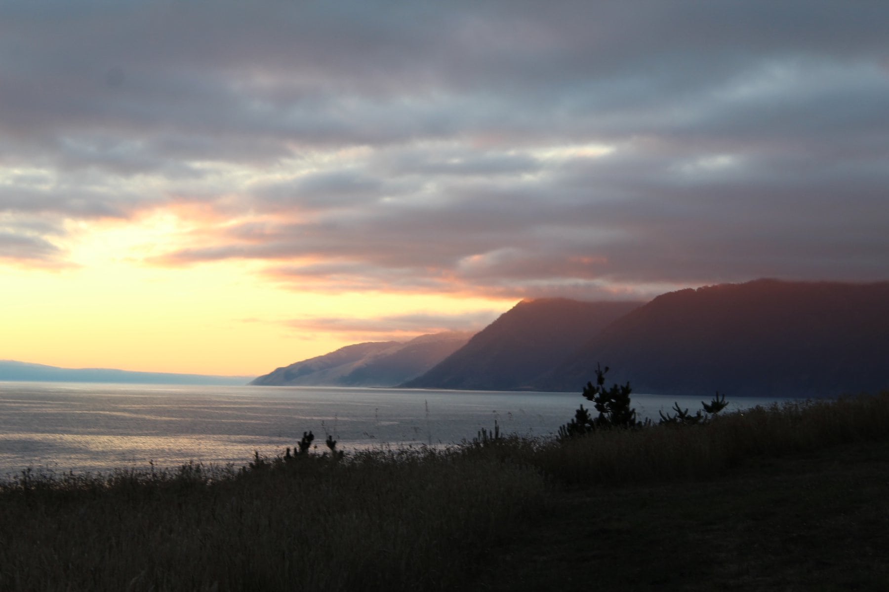 A serene coastal scene at sunset with mountains in the background partially covered by clouds, and the calm sea reflecting the warm hues of the sky.