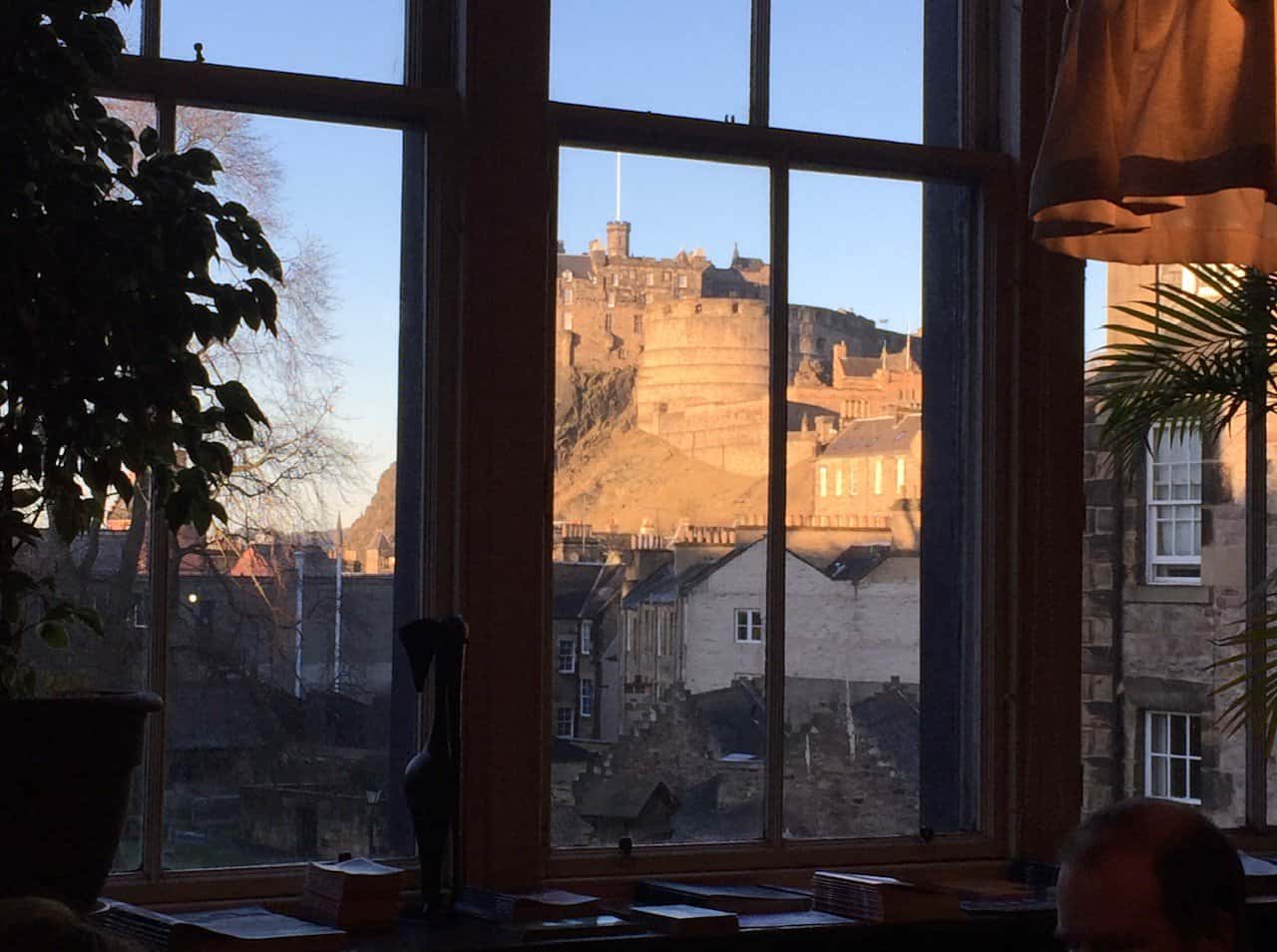 View of Edinburgh Castle from inside a room, framed by a window with a plant on the left. The castle is bathed in sunlight, highlighting its stone walls and the surrounding rooftops.