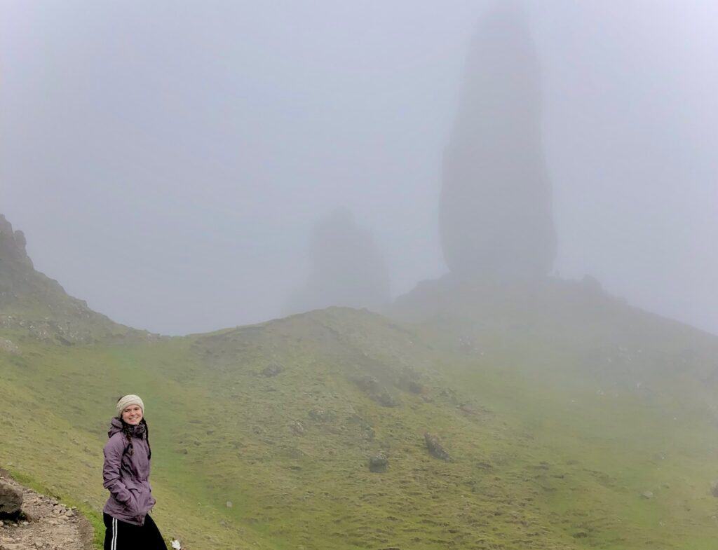 A woman wearing a purple jacket and a beige headband stands smiling in front of a misty, grassy Isle of Skye with large rock formations called the Old Man of Storr partially obscured by fog.  