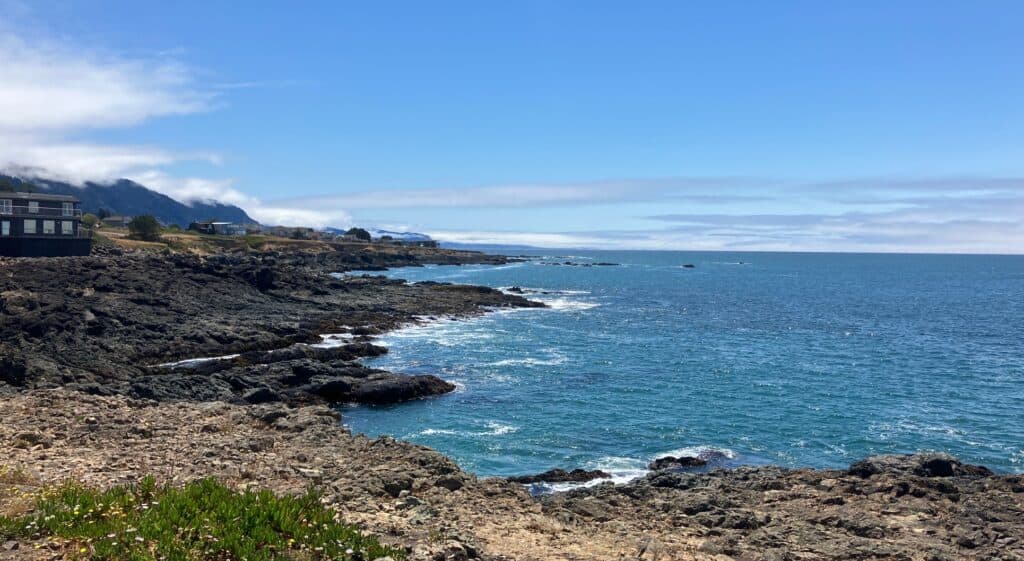 Rocky coastline with clear blue ocean water, Shelter Cove houses along the shore, and mountains in the background partially covered by clouds. The sky is bright and mostly clear.