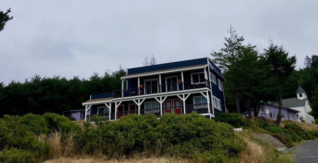 A two-story navy blue house with white trim and red doors, situated on a hillside with greenery in the foreground. The sky is overcast, and a smaller building is visible to the right, partially obscured by trees.