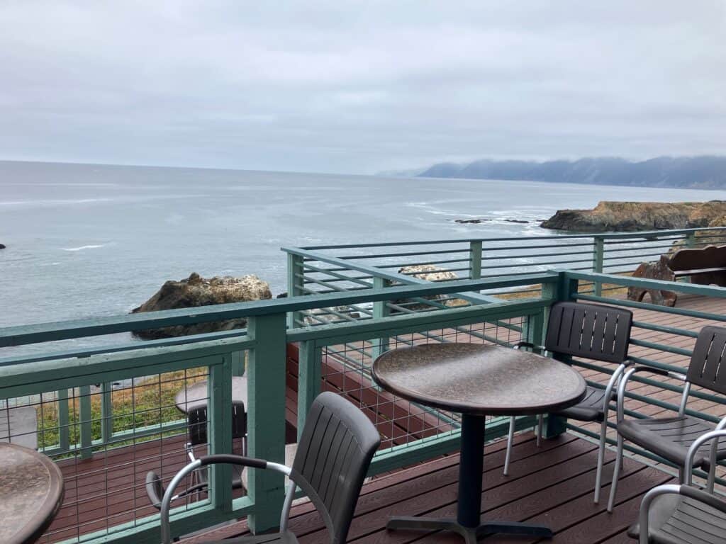 Outdoor dining table with a blue railing surrounding the deck. Overlooks the ocean waters and mountains in the distance of Shelter Cove.