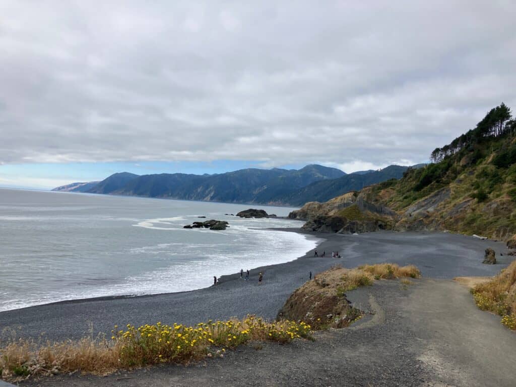 A rocky coastline with dark sand and a few people walking along the shore. The ocean waves are gently rolling in at the Black Sands Beach in Shelter Cove, and the coastline stretches towards forested hills in the distance under a cloudy sky.