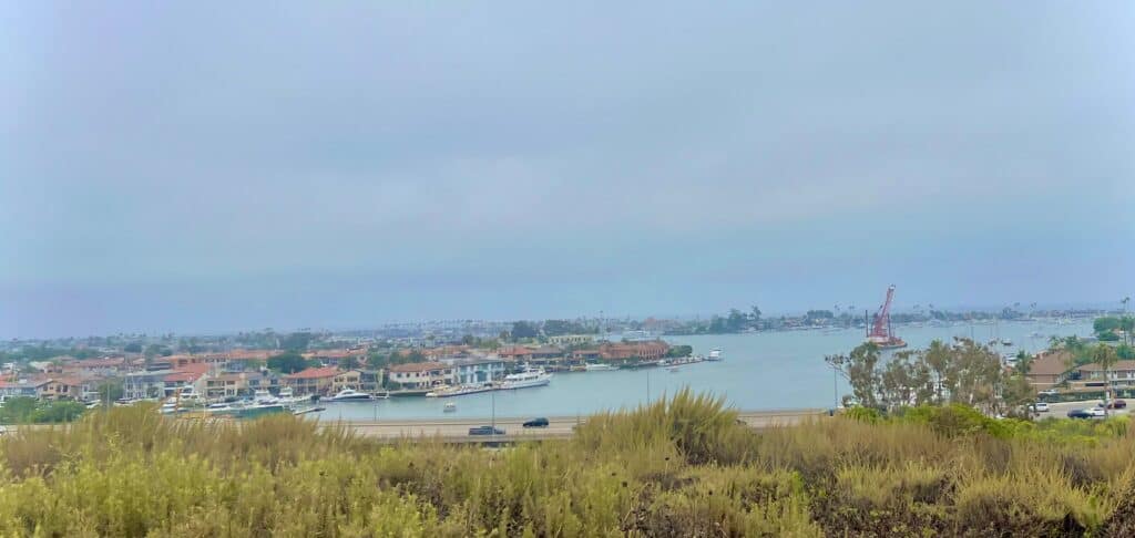 A view of a coastal town with houses and buildings along the shoreline, a body of water with boats docked, and a large construction crane in the distance. The sky is overcast, casting a soft light over the scene.