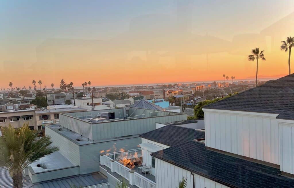 View of a coastal town at sunset, with palm trees and rooftops silhouetted against an orange and blue sky. The foreground shows modern buildings, while the distant horizon meets the ocean.