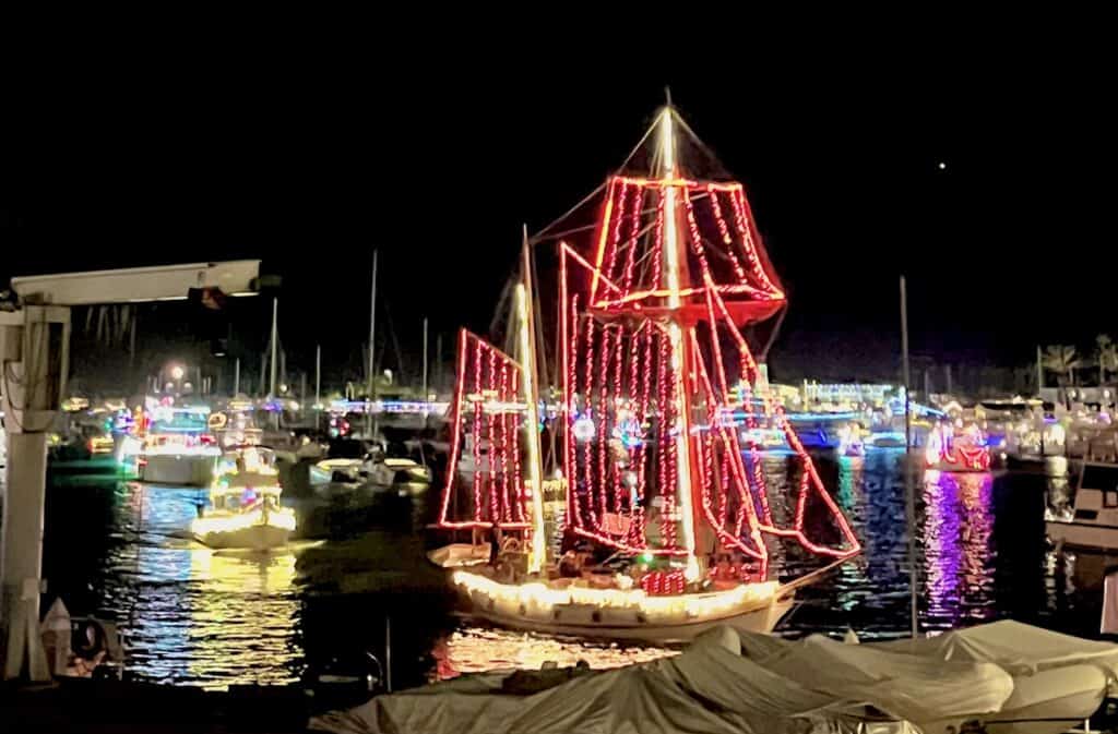 Boats adorned with vibrant Christmas lights docked at a marina during nighttime. The central boat is decorated to resemble a tall ship with red lights outlining its sails.