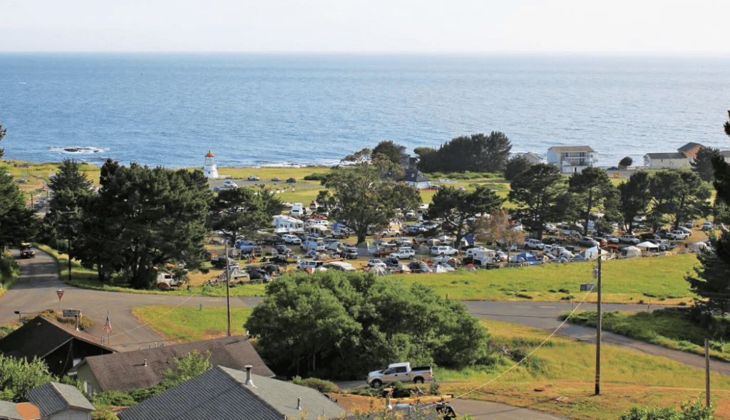 A coastal town with a lighthouse, numerous parked cars, and tents, indicating a bustling camping area near the ocean on a sunny day.