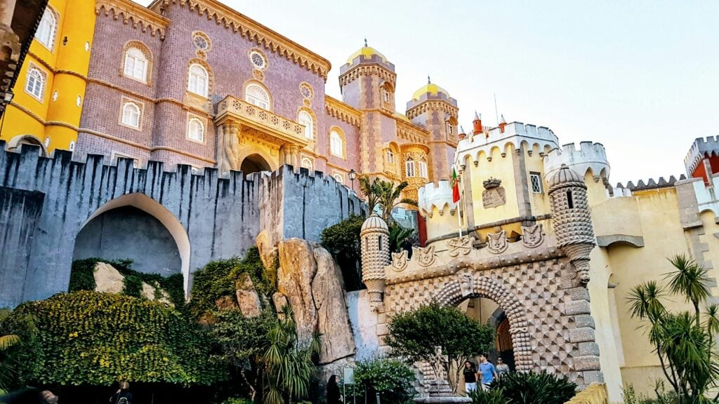 A vibrant photo of Pena Palace in Sintra, Portugal, showcasing its eclectic architectural styles with colorful facades, decorative battlements, and lush vegetation around the castle.