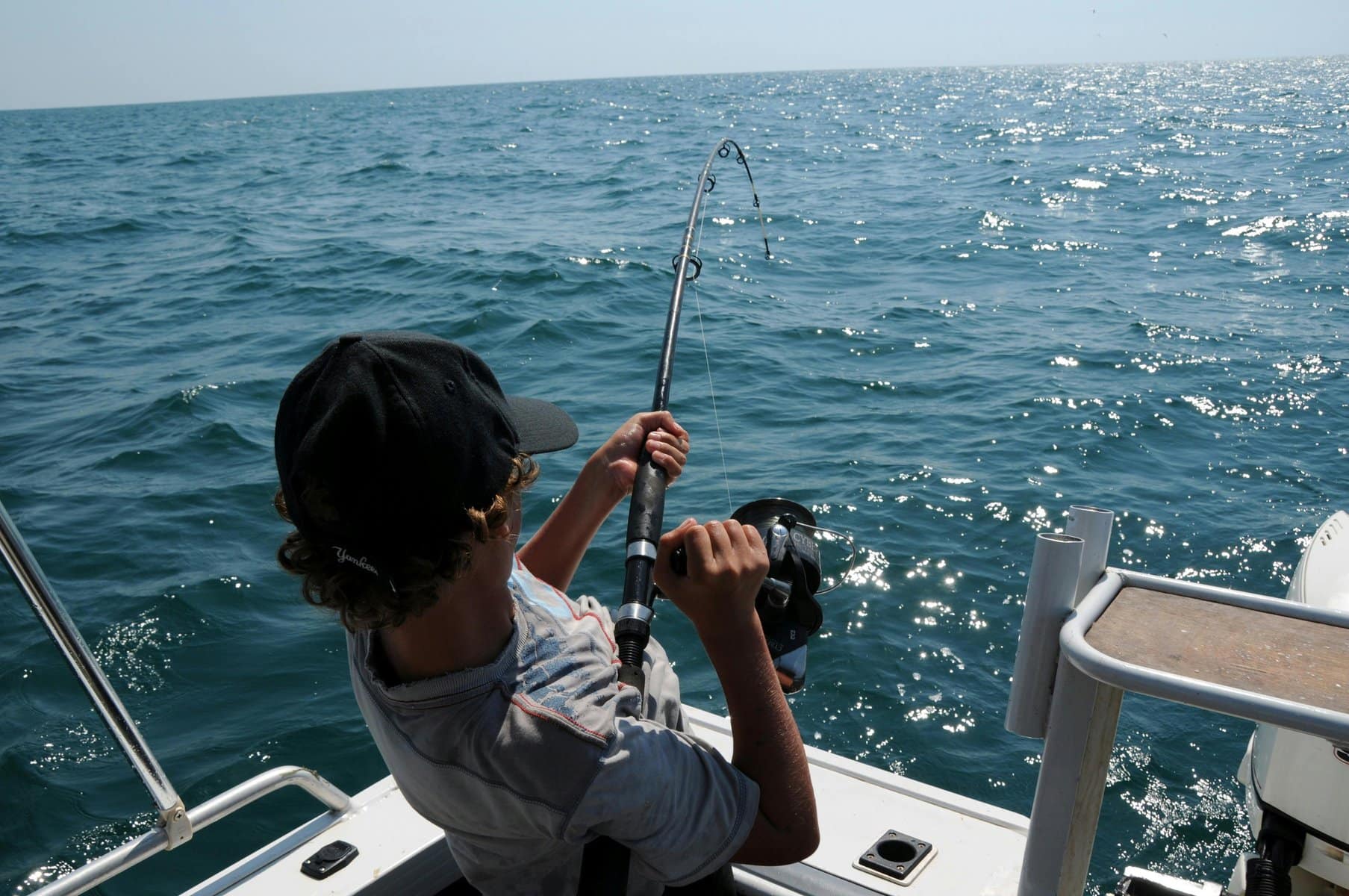 Man with baseball hat reels in fishing pole in the ocean during the daytime.